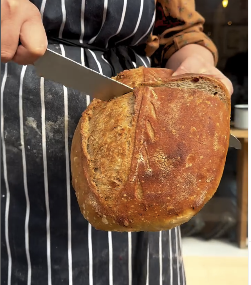 Slice of sourdough being held in a left hand and sliced using a bread knife held in the right hand