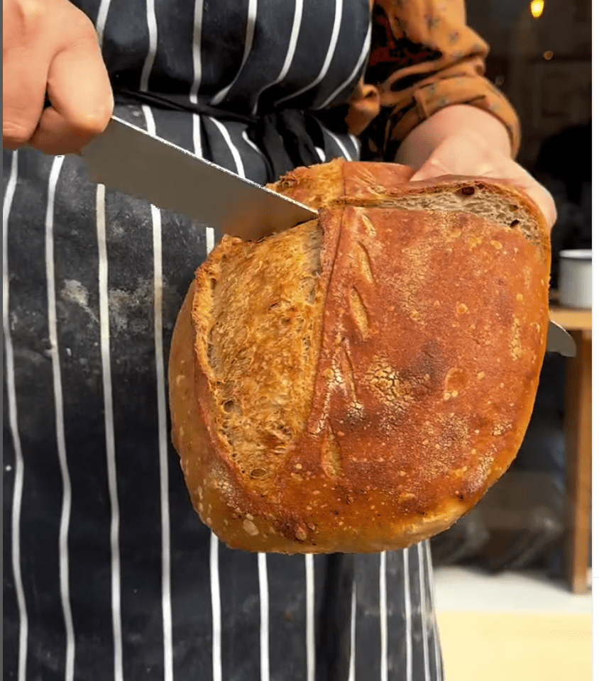 Slice of sourdough being held in a left hand and sliced using a bread knife held in the right hand